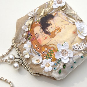 Puede incluir: Un bolso de mano de terciopelo beige con cierre dorado y una cadena de perlas. El bolso est&aacute; decorado con flores blancas, cuentas verdes y una imagen impresa de una pintura de dos mujeres abraz&aacute;ndose.