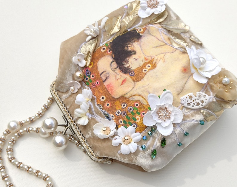 Puede incluir: Un bolso de mano de terciopelo beige con cierre dorado y una cadena de perlas. El bolso est&aacute; decorado con flores blancas, cuentas verdes y una imagen impresa de una pintura de dos mujeres abraz&aacute;ndose.