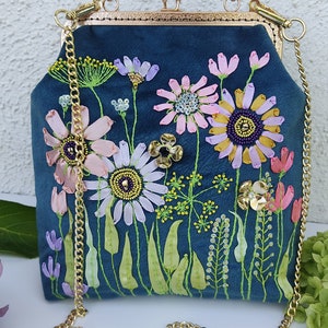 K&ouml;nnte beinhalten: Eine blaue Samt-Handtasche mit einer goldenen Kettenriemen und einem gestickten Blumenmuster. Die Blumen sind rosa, wei&szlig; und gelb mit gr&uuml;nen Bl&auml;ttern und Stielen.