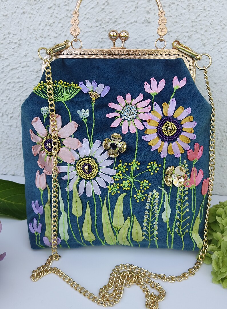 K&ouml;nnte beinhalten: Eine blaue Samt-Handtasche mit einer goldenen Kettenriemen und einem gestickten Blumenmuster. Die Blumen sind rosa, wei&szlig; und gelb mit gr&uuml;nen Bl&auml;ttern und Stielen.