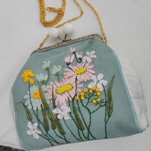 May include: A light blue satin clutch bag with a gold chain strap and embroidered floral design. The flowers are pink, white, and yellow.