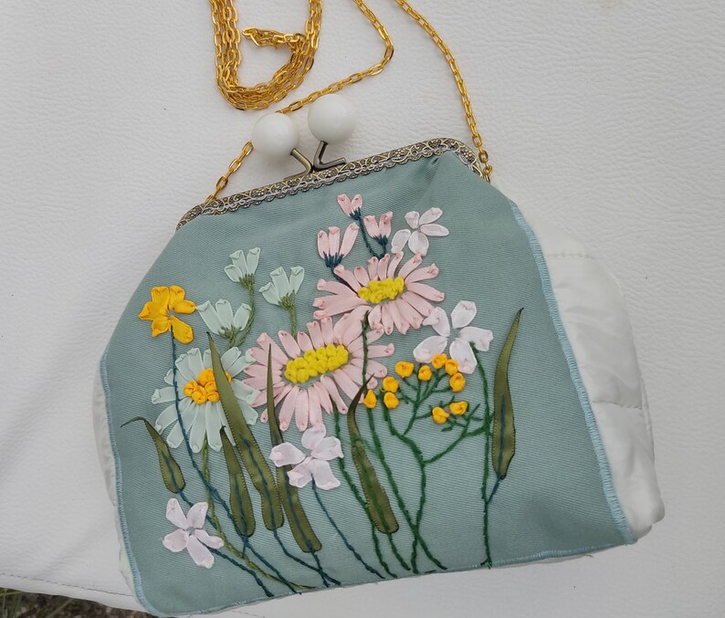 May include: A light blue satin clutch bag with a gold chain strap and embroidered floral design. The flowers are pink, white, and yellow.