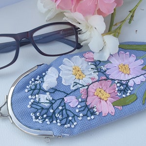 May include: A light blue eyeglass case with a floral embroidery design. The case has a silver metal clasp and is decorated with pink, white, and green flowers.