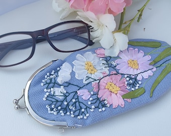 May include: A light blue eyeglass case with a floral embroidery design. The case has a silver metal clasp and is decorated with pink, white, and green flowers.