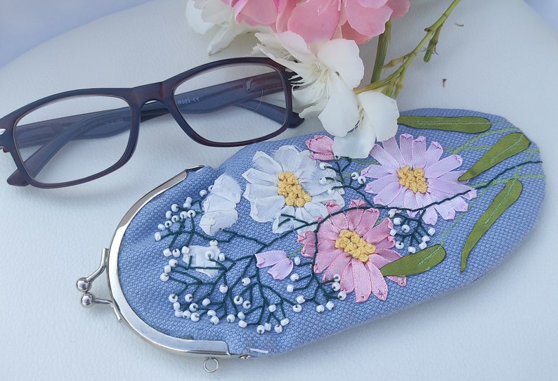 May include: A light blue eyeglass case with a floral embroidery design. The case has a silver metal clasp and is decorated with pink, white, and green flowers.