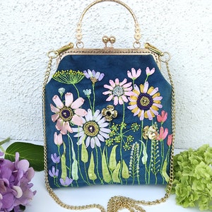 K&ouml;nnte beinhalten: Eine blaue Samt-Handtasche mit einer goldenen Kettenkette und einem gestickten Blumendesign. Die Blumen sind rosa, wei&szlig; und gelb mit gr&uuml;nen Bl&auml;ttern.