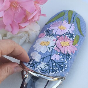 May include: A light blue fabric glasses case with a floral embroidery design. The case has a silver metal clasp and is decorated with pink and white flowers, green leaves, and white beads.