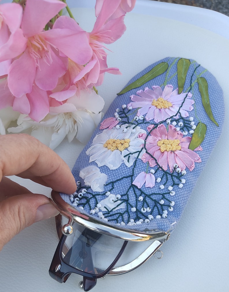 May include: A light blue fabric glasses case with a floral embroidery design. The case has a silver metal clasp and is decorated with pink and white flowers, green leaves, and white beads.
