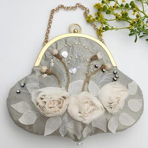 May include: A gray, embellished handbag with a gold chain handle. The bag features white fabric flowers, beaded accents, and a decorative clasp. The bag is approximately 10 inches wide.