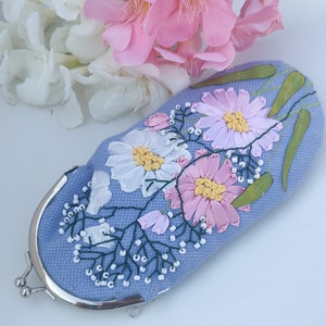 May include: A light blue fabric purse with a floral design embroidered in pink, white, and green. The purse has a silver clasp and a silver chain.