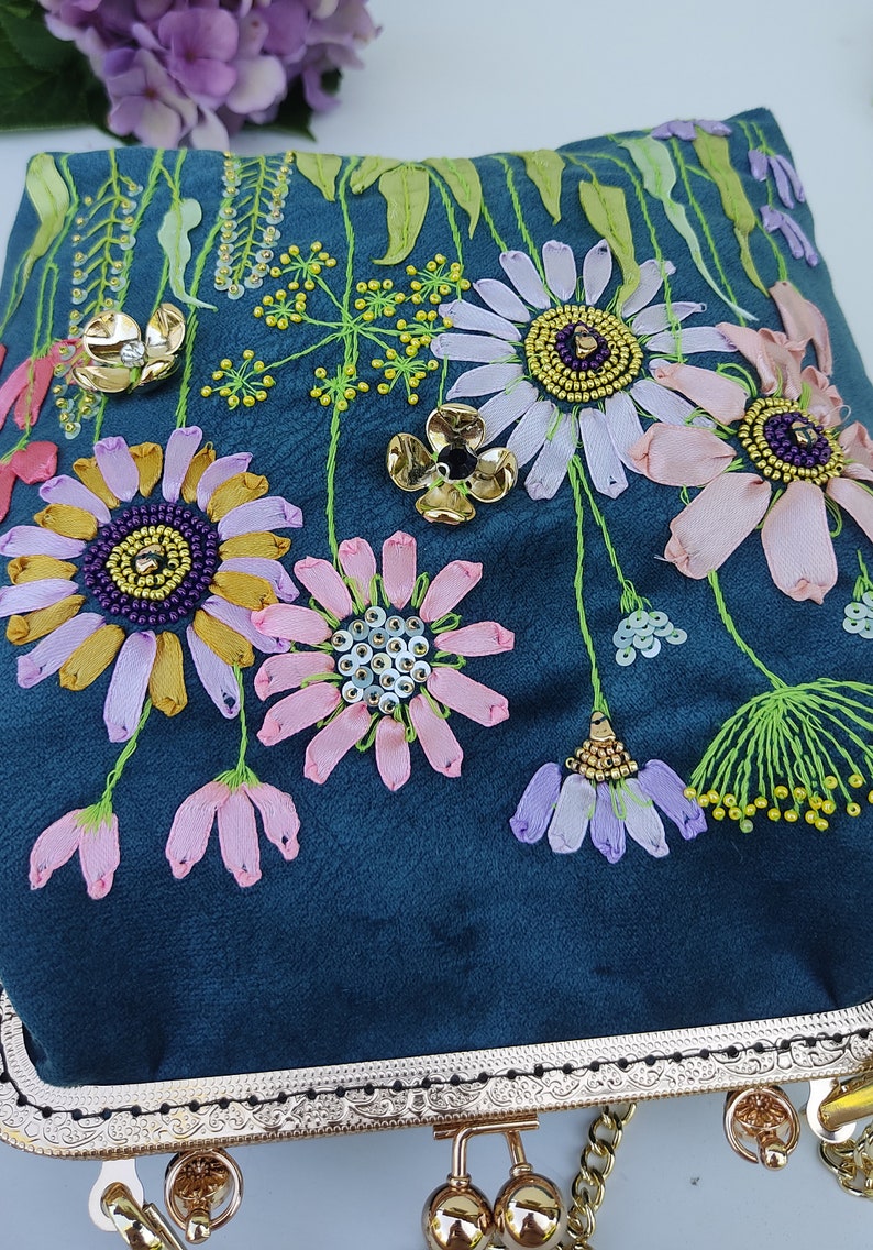 K&ouml;nnte beinhalten: Eine Clutch aus blaugr&uuml;nem Samt mit goldenem Metallrahmen und Kette. Die Clutch ist mit bunten Blumen und Perlen bestickt.