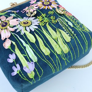 K&ouml;nnte beinhalten: Eine tealblaue Samt-Handtasche mit einer goldenen Kettenkette. Die Tasche ist mit gestickten Blumen und Bl&auml;ttern in den Farben Rosa, Lila, Gr&uuml;n und Gelb verziert.