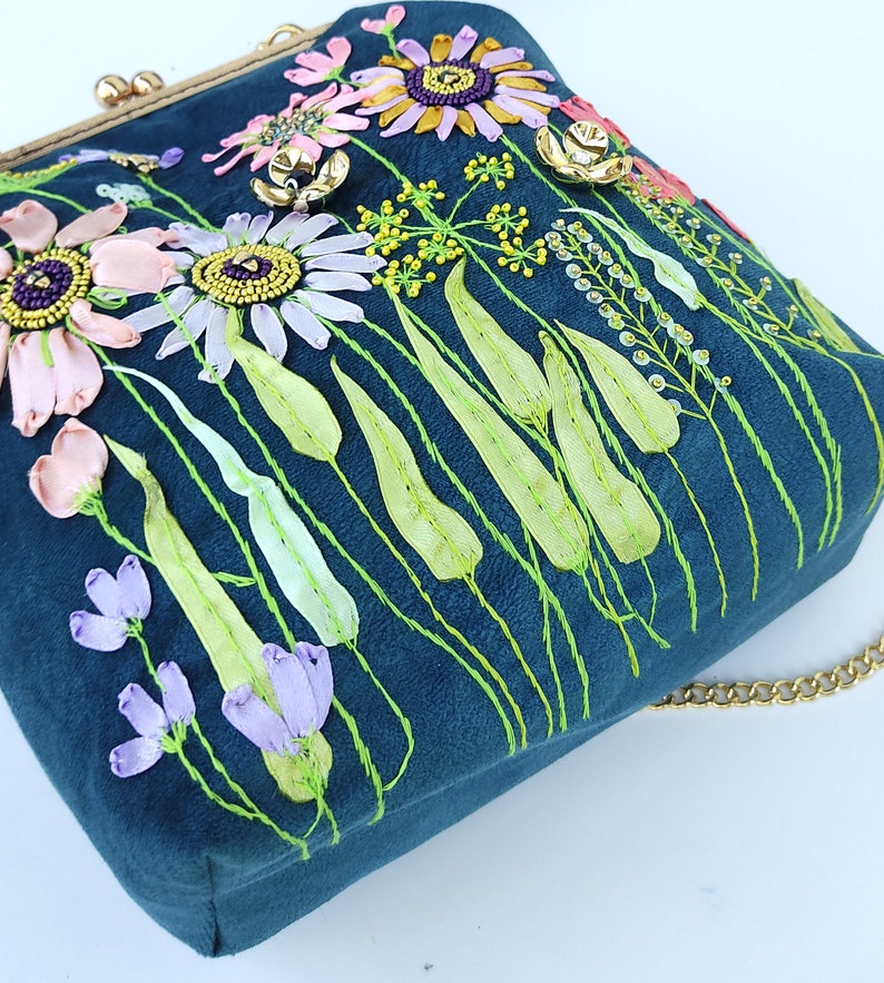 K&ouml;nnte beinhalten: Eine tealblaue Samt-Handtasche mit einer goldenen Kettenkette. Die Tasche ist mit gestickten Blumen und Bl&auml;ttern in den Farben Rosa, Lila, Gr&uuml;n und Gelb verziert.