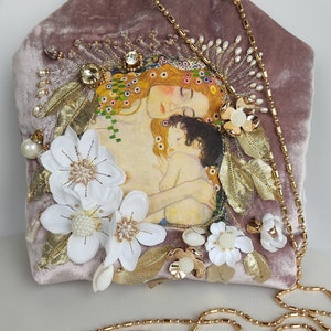 Puede incluir: Un bolso de mano de terciopelo malva con una cadena de oro. El bolso est&aacute; decorado con flores blancas, hojas doradas y una imagen impresa de una mujer y un ni&ntilde;o.