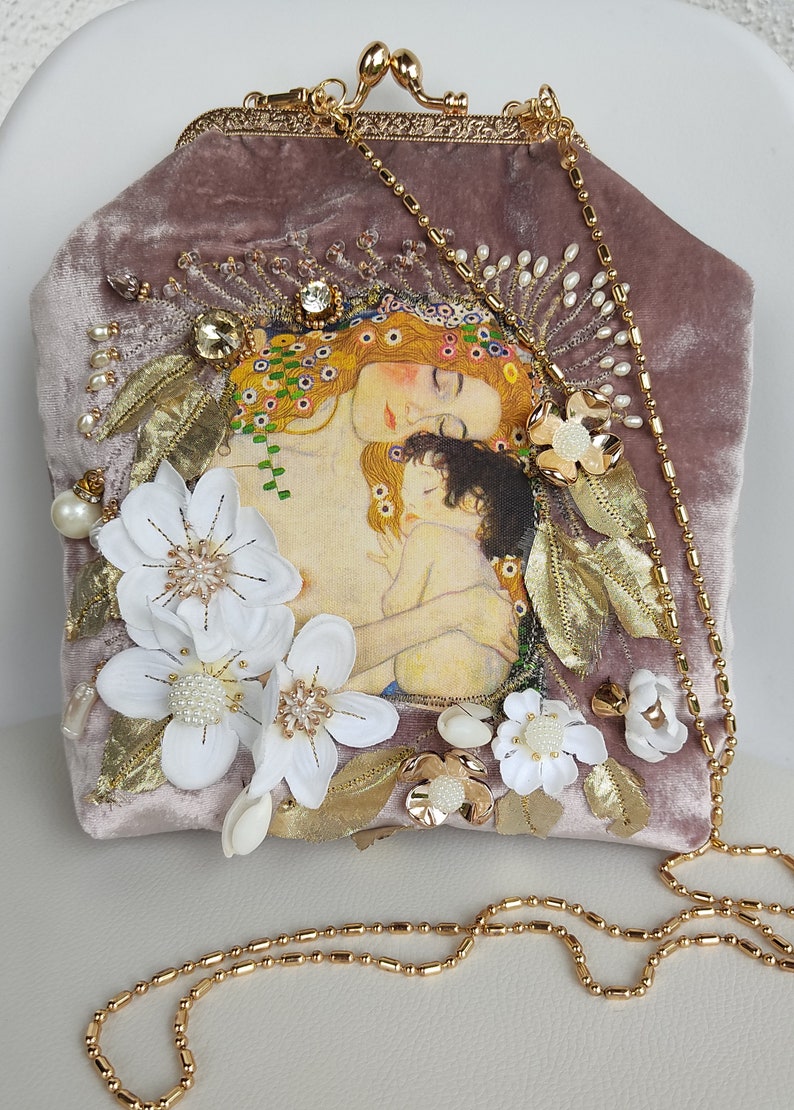 Puede incluir: Un bolso de mano de terciopelo malva con una cadena de oro. El bolso est&aacute; decorado con flores blancas, hojas doradas y una imagen impresa de una mujer y un ni&ntilde;o.