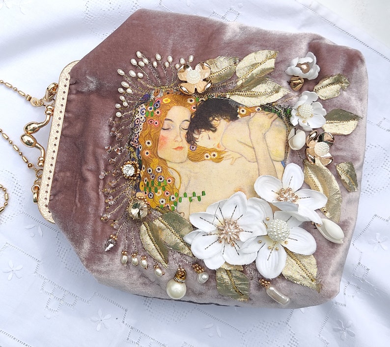 Puede incluir: Una cartera de mano de terciopelo malva con una cadena de oro y un dise&ntilde;o de cuentas que representa una pintura de dos figuras abraz&aacute;ndose. La cartera est&aacute; decorada con flores blancas y detalles dorados.
