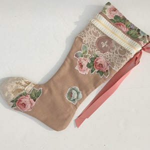 Può includere: Una calza di Natale beige con applicazioni di rose floreali e finiture in pizzo. La calza presenta un nastro rosa, un emblema del giglio e un polsino fantasia con rose rosa e verdi. La punta è in tessuto color crema.