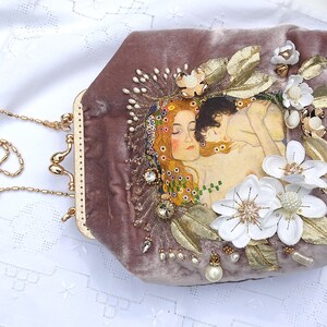 Puede incluir: Un bolso de mano de terciopelo malva con una cadena de oro y un dise&ntilde;o floral. El dise&ntilde;o presenta flores blancas, hojas doradas y una pintura de dos mujeres.