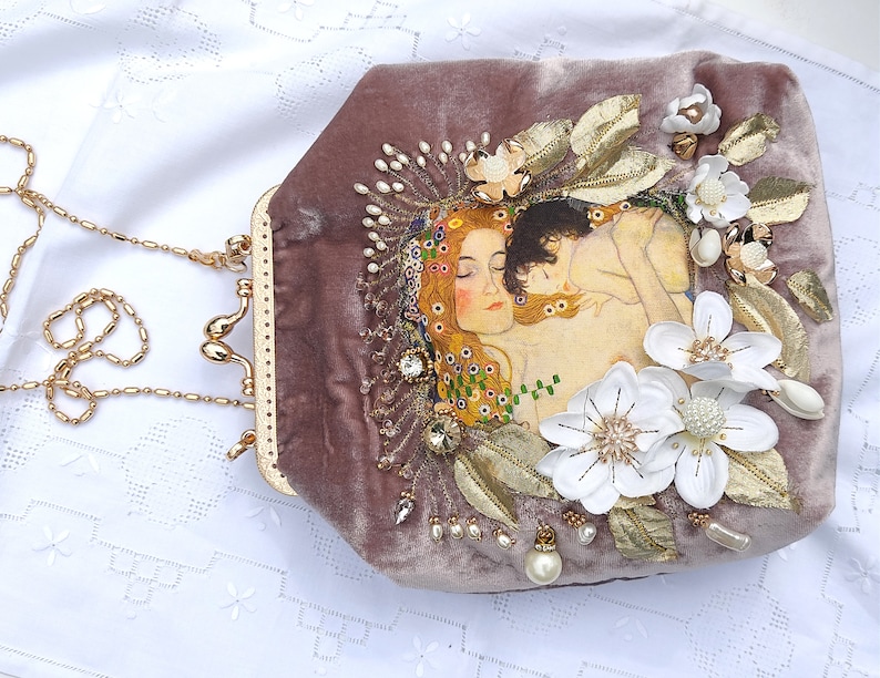 Puede incluir: Un bolso de mano de terciopelo malva con una cadena de oro y un dise&ntilde;o floral. El dise&ntilde;o presenta flores blancas, hojas doradas y una pintura de dos mujeres.