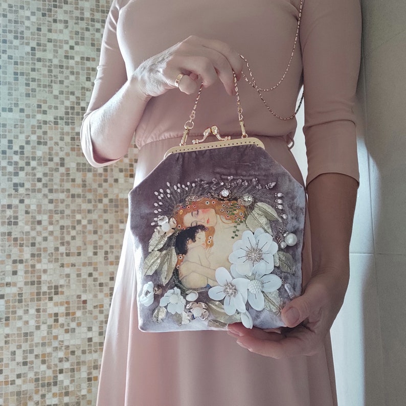 Puede incluir: Un bolso de mano de terciopelo morado con cierre de broche dorado y correa de cadena. El bolso presenta una imagen bordada de dos mujeres con flores y un dise&ntilde;o floral.