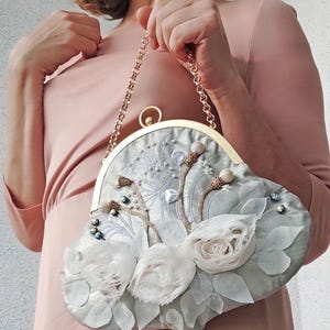 May include: A light blue clutch purse with a gold chain strap. The purse is decorated with white fabric flowers, leaves, and beaded accents. The clasp and chain are gold-colored.