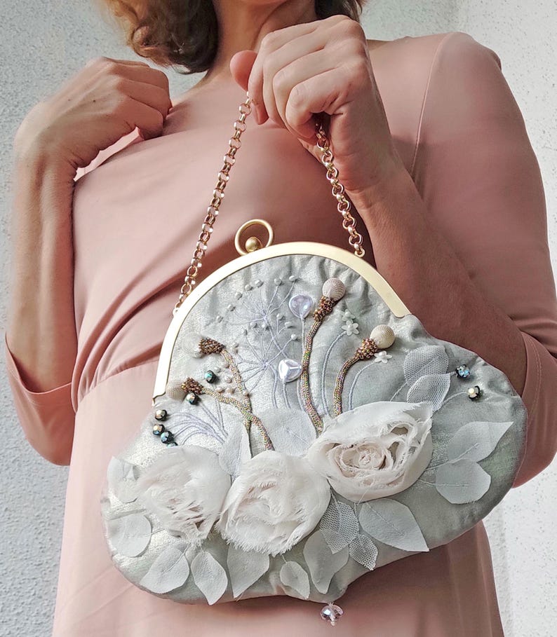 May include: A light blue clutch purse with a gold chain strap. The purse is decorated with white fabric flowers, leaves, and beaded accents. The clasp and chain are gold-colored.
