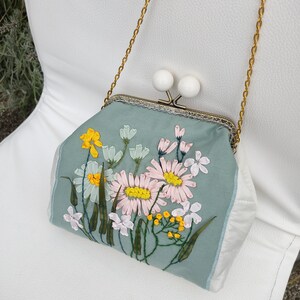 May include: A light blue fabric purse with a floral embroidery design. The purse has a gold chain strap and a clasp closure with two white decorative knobs.