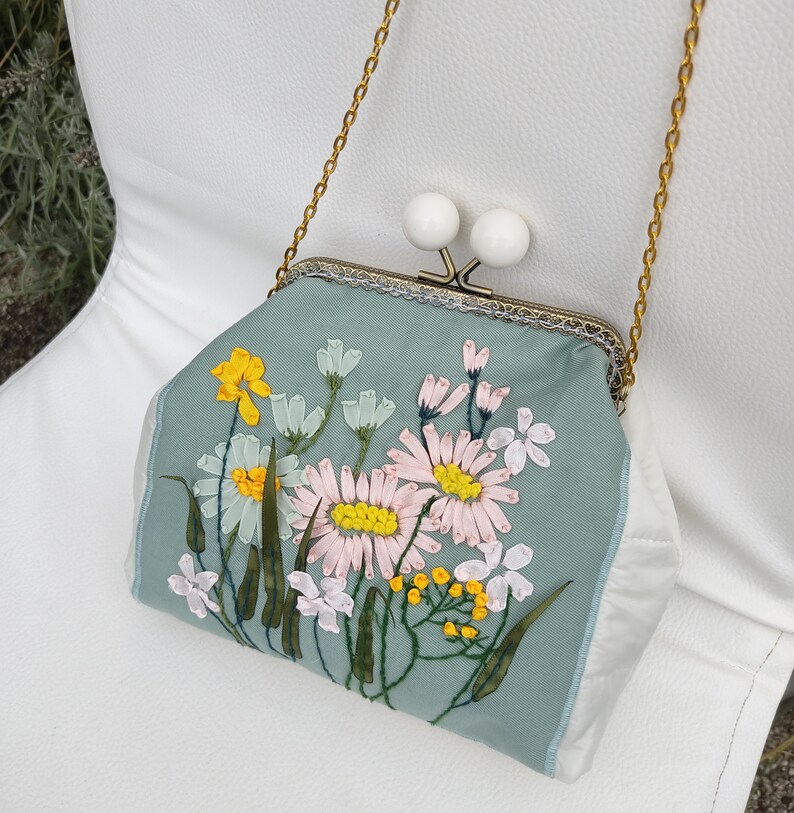 May include: A light blue fabric purse with a floral embroidery design. The purse has a gold chain strap and a clasp closure with two white decorative knobs.