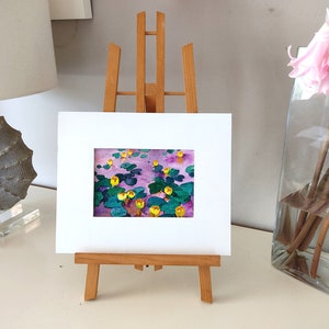 May include: A framed painting of yellow water lilies on a purple background, displayed on a small wooden easel.