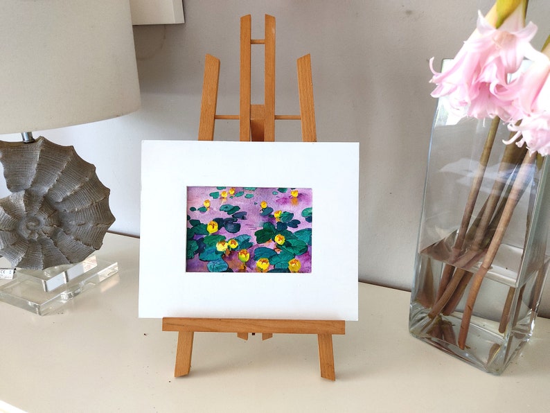 May include: A framed painting of yellow water lilies on a purple background, displayed on a small wooden easel.