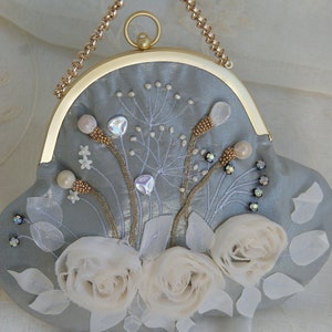 May include: A light blue clutch with a gold-tone frame and chain handle. The clutch is embellished with white fabric roses, embroidered floral details, and sparkling crystals. The bag measures approximately 8 inches wide.