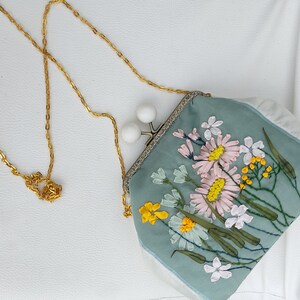 May include: A light blue fabric purse with a floral embroidery design. The purse has a gold chain strap and a white clasp.