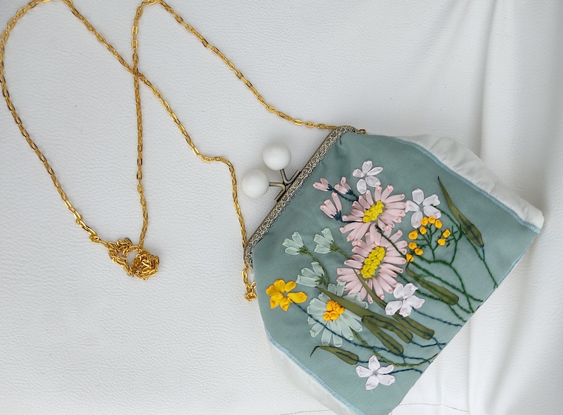 May include: A light blue fabric purse with a floral embroidery design. The purse has a gold chain strap and a white clasp.