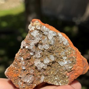 May include: A natural mineral specimen featuring a rough, orange-red exterior with a cluster of clear, crystalline formations. The crystals are clustered on a brown and tan base, creating a striking contrast. The specimen is held in a hand.