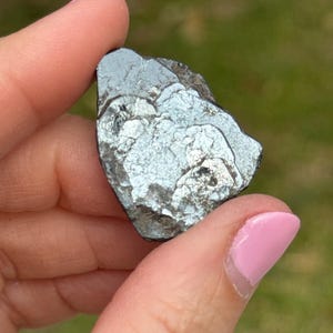 May include: A dark, metallic-looking mineral specimen with a layered, textured surface. The stone has a silver-gray color with reflective qualities. The edges are rough and uneven, suggesting a natural, unpolished state. The background is blurred green.