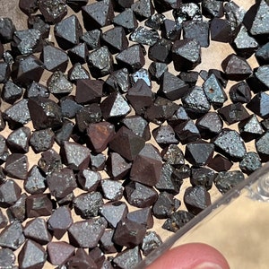 Pode incluir: Um close-up de um recipiente cheio de pequenos cristais octaédricos metálicos de cor cinza escuro. Os cristais estão dispostos em um padrão aleatório e têm uma superfície brilhante e reflexiva.