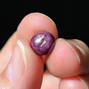 May include: A deep red ruby gemstone with a star-like pattern in the center. The gemstone is faceted and has a smooth, polished surface.