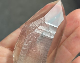 May include: A clear quartz crystal with a pointed tip and a rough, natural base. The crystal has a light brown hue and is held in a hand. The crystal is a natural mineral specimen.