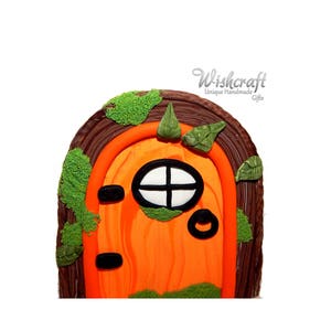 Puede incluir: Puerta de hadas naranja con marco de madera marrón, una pequeña ventana y musgo verde alrededor de los bordes. La puerta está decorada con una perilla de puerta negra y una línea negra en el centro. El texto "Wishcraft Unique Handmade Gifts" es visible en la esquina superior derecha de la imagen.