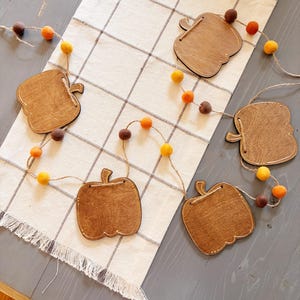 May include: A decorative garland featuring five wooden pumpkin cutouts strung together with twine. The garland is accented with small felt balls in shades of orange, yellow, and dark brown, spaced evenly between the wooden pumpkin shapes.