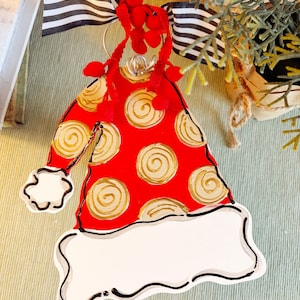 May include: A red and white Santa hat-shaped Christmas ornament. The hat features gold swirl designs on the red portion and a white brim. It is adorned with red pom-poms and a black and white striped bow. A festive holiday decoration.