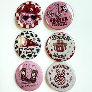 May include: Six circular pin-back buttons featuring University of Oklahoma themes. Designs include a disco ball with a cowboy hat, cherries, a cow print flag with 'always a sooner', a football with 'Boomer Baby', cowboy boots, and a peace sign.