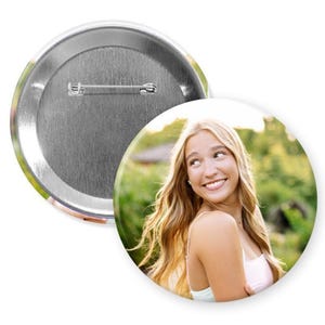 May include: A circular pin-back button featuring a photographic portrait of a smiling person with long blonde hair. The reverse side of the button is metallic silver and includes a horizontal pin mechanism for attachment to clothing or fabric items.
