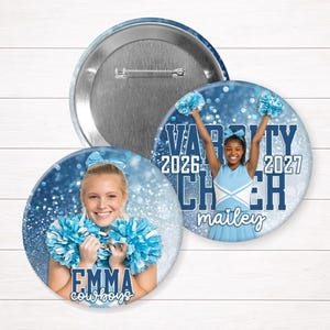 May include: Two round pinback buttons featuring cheerleaders holding blue pom-poms against a blue glitter background. One button shows a cheerleader named Emma, the other shows a cheerleader named Mailey with the text Varsity Cheer 2026 2027.