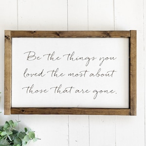 May include: A rectangular wooden sign with a dark brown stained frame and a white center panel. The sign features black cursive text that reads: Be the things you loved the most about those that are gone. The sign has a clean, simple design.