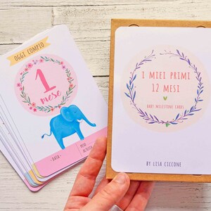 May include: A set of 12 baby milestone cards with a floral wreath design. The cards are in a brown box and feature a blue elephant illustration on the front. The text on the box reads "I MIEI PRIMI 12 MESI BABY MILESTONE CARDS BY LISA CICCONE".