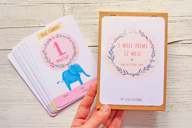 May include: A set of 12 baby milestone cards with a floral wreath design. The cards are in a brown box and feature a blue elephant illustration on the front. The text on the box reads "I MIEI PRIMI 12 MESI BABY MILESTONE CARDS BY LISA CICCONE".
