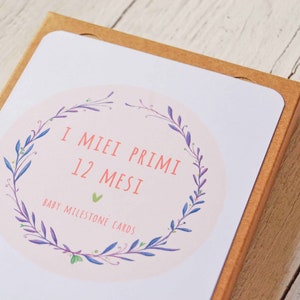 May include: A box of baby milestone cards with a floral wreath design. The box is brown and the cards are white with pink and green accents. The text on the box reads "I Miei Primi 12 Mesi Baby Milestone Cards".