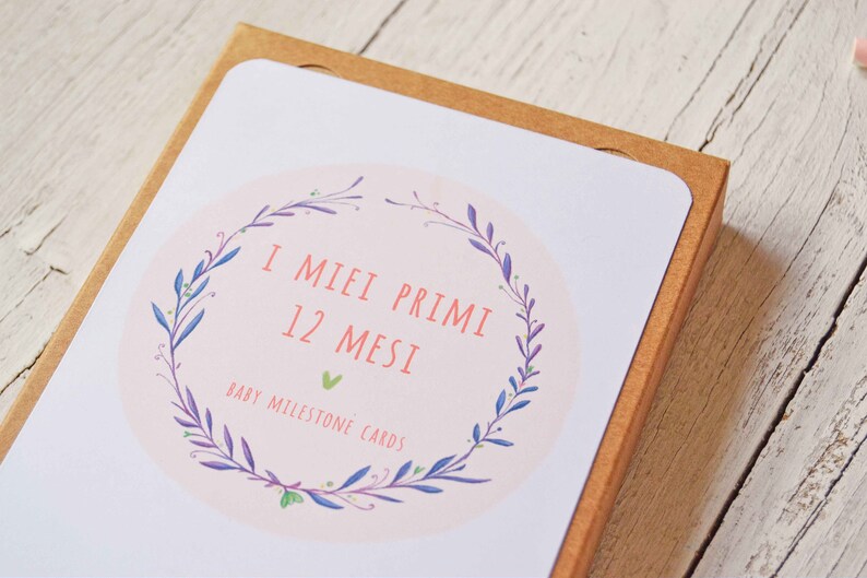 May include: A box of baby milestone cards with a floral wreath design. The box is brown and the cards are white with pink and green accents. The text on the box reads "I Miei Primi 12 Mesi Baby Milestone Cards".