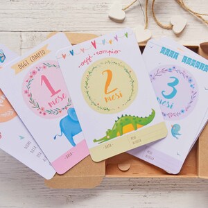 May include: Four colorful baby milestone cards with illustrations of an elephant, crocodile, and bird. The cards are in a cardboard box and feature the text "Oggi compio" and numbers 1, 2, and 3.